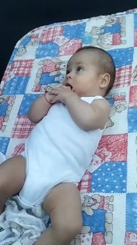 In the bright daylight of Fălești, Moldova, a baby lies on a colorful quilt, its white onesie contrasting with the blanket's playful teddy bear pattern. The baby, with light skin and dark hair, appears to be resting, with its hands near its mouth. The quilt is laid out on a dark surface, likely a trampoline, suggested by the texture and the presence of a child's hands appearing in the frame, clapping.

The camera then shifts to focus on a young girl, adorned in a pink lace dress, with blonde hair pulled up. She is on the trampoline, her face partially obscured by her hand as she looks towards the camera. In a subsequent frame, she lowers her hand, revealing her blue eyes and a serious expression. The background shows a fence and lush greenery, indicating an outdoor setting. The overall atmosphere is one of outdoor play and childhood activity on a pleasant day. There is no visible text in the media.