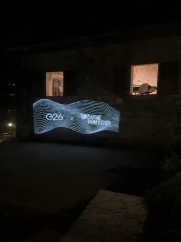 A video captures a nighttime scene of a stone building with two windows. A projector casts a white, wavy graphic with text onto the wall to the left of the windows. The text is partially obscured by the wavy lines, but "26" and what appears to be "WHITE" and "WATER" are visible. The area in front of the building is dark, with some stone paving. To the right of the building, a pathway leads into darkness, illuminated by a distant light source and some ambient light from the windows. The windows themselves reveal dimly lit interiors. The video is brief, with a slight panning motion to the right in the middle.