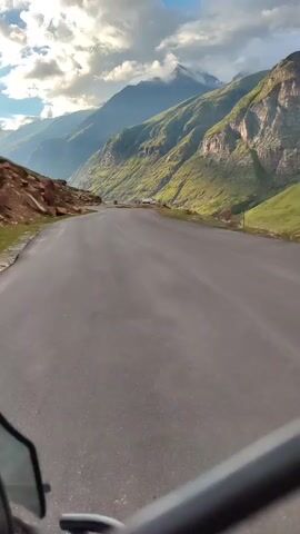 A first-person perspective video shows a paved road winding through a mountain valley. Green grassy slopes and rugged, rocky mountain peaks dominate the landscape. The sky is partly cloudy, with sunlight breaking through. The road appears empty.
