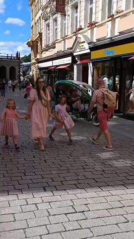 The video shows a street scene with pedestrians and a three-wheeled electric rickshaw. In the foreground, a person wearing a bright pink jacket is facing away from the camera. A young girl in a light-colored dress and a man with a backpack are walking to the left. In the middle ground, a green electric rickshaw is moving to the right. The rickshaw has a canopy and seats two passengers. A man is driving the rickshaw. Behind the rickshaw, people are walking on the sidewalk. The background consists of buildings with storefronts. The street is paved with cobblestones.