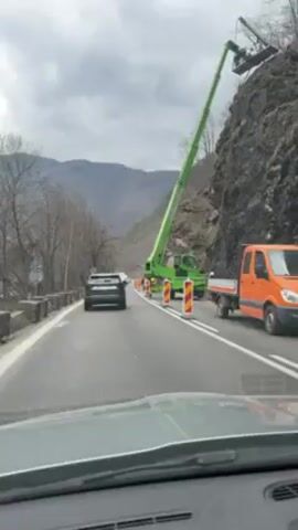 A car drives along a road that runs parallel to a steep rock face. A large green construction crane is positioned along the road with its boom extended high up the rock face, where two people appear to be working from a platform. Traffic cones are set out to direct traffic around the work area. An orange pickup truck is parked on the shoulder of the road. The setting is a mountainous area with trees in the background and an overcast sky.