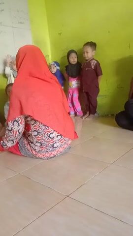 A group of young children, appearing to be of preschool or early elementary age, are arranged in a line on the floor. They are facing away from the camera. One adult, presumably a teacher or caregiver, is kneeling down in front of them, facing the children. This adult is wearing a bright red headscarf and a patterned dress. In their hands, the adult appears to be holding a white container and a small object, possibly a treat or prize, which they are offering to the children. The children are dressed in various colorful outfits, some wearing hijabs. The floor is tiled, and the wall behind the children is painted a bright green. There is some writing or drawings on the green wall. The overall impression is of an educational or recreational setting, possibly a classroom or a learning activity. The video appears to be taken from a low angle, possibly by another child or adult in the room. The image is slightly tilted.