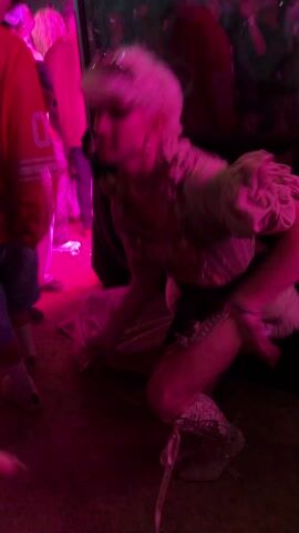 The video captures a performance on a raised platform in what appears to be a crowded outdoor event, possibly a festival. A person dressed in a peculiar costume that includes a bodice, shorts, and ruffled leg warmers is dancing. The person is also wearing a white wig. A mirror on the platform reflects the crowd, including a person wearing an orange jersey. The lighting is dominated by pink hues, creating a vibrant atmosphere. A hand holding a beverage is visible in the foreground, implying the videographer is part of the audience enjoying the performance.