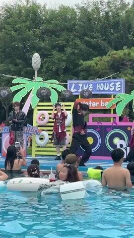 The video shows an outdoor performance at a pool party. Three young men, dressed in stylish, modern clothing, are performing on a stage decorated with palm trees, inflatable rings, and a boombox graphic. The man on the right is wearing a black and red checkered jacket and black pants, while the man in the middle wears a red and black checkered shirt and grey shorts, and the man on the left wears a black jacket and grey cargo pants. They appear to be dancing and singing. In the foreground, several people are enjoying the pool, some of them wearing inflatable rings. They are watching the performance. The background features lush green trees and a cloudy sky. The overall atmosphere is festive and energetic.