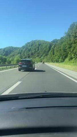 This is a first-person perspective video of driving on a highway. The camera is positioned inside a car, looking forward. The highway has multiple lanes, marked by white lines. On the left side of the highway, there's a barrier and then a grassy area with trees. On the right side, there are also trees and a grassy shoulder. In the distance, there are rolling hills covered in dense green foliage. The sky is a clear, bright blue.

There are several cars visible on the highway. In the foreground, the dashboard of the car is visible. Further ahead, a dark-colored SUV is in the right lane. Another lighter-colored car is in the left lane. Even further ahead, there's a yellow vehicle, possibly a truck or a van. The cars are moving in the same direction as the viewer's car. The overall impression is of a sunny day driving through a scenic, mountainous area.