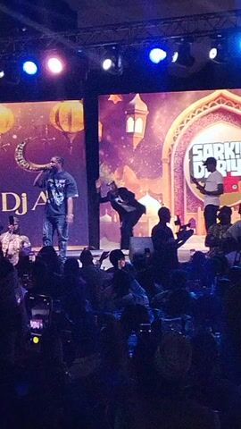 The scene captures a live performance on a brightly lit stage in front of a large audience. The backdrop features two large screens with Arabic-themed graphics and the text "DJ AB" on one and "SARKIN DARIYA MULTIPLE LAFFS" on the other. There are three men on stage. One man in a dark t-shirt and jeans holds a microphone and appears to be rapping or singing. Another man in a suit is dancing energetically. The third man in a white t-shirt and jeans stands to the side, appearing to hold a phone. The audience is seated and many are holding up their phones to record the performance. The lighting is colorful with spotlights aimed at the stage. The overall environment suggests a concert or live entertainment event with an audience enjoying the show.