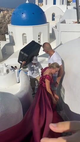 A man wearing a white t-shirt and a baseball cap carries a large black suitcase on his right shoulder. He is walking up a set of white stairs. Beside him, a woman in a flowing burgundy dress is being helped down the stairs by a man in a white t-shirt and blue shorts. In the background, a white building with blue domes and crosses is visible, overlooking the sea. The environment appears to be a sunny, coastal location, possibly a Greek island, with boats visible on the water.