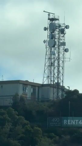 The video shows a tall telecommunications tower situated on a hill next to a building. The tower is a complex structure with numerous antennas and dishes of various sizes, including a large radar dish at the very top. The building appears to be a utilitarian structure, possibly an operations center for the tower, with windows and some visible equipment. Lush green trees surround the base of the hill, and a billboard is visible in the lower right corner of the frame, partially obscured by foliage, with text that includes "490 AVENUE OF THE PALMS" and "RENT490.COM". The sky is overcast, contributing to a muted lighting condition. No people are visible in the video, and no actions or interactions are depicted. The camera appears to be static, providing a consistent view of the scene.