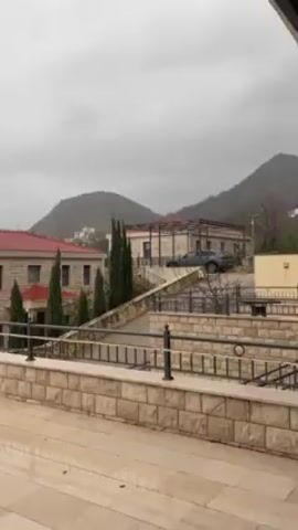 The video shows an outdoor scene, likely a patio or terrace, on an overcast day. The surface of the patio is wet, suggesting it has recently rained. There are tables and chairs present, indicating a space for leisure or dining. In the background, there are stone buildings with red-tiled roofs and meticulously landscaped greenery including tall, thin trees. A mountain range is visible in the distance, partially obscured by the gray, cloudy sky. A car is parked on a sloped driveway behind one of the buildings. The camera slowly pans across the scene.