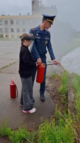 In an outdoor setting in Markovskii, Russia, a young girl is being instructed by a man in a dark blue uniform on how to use a fire extinguisher. The girl, wearing a headset or visor, holds a red fire extinguisher and aims the nozzle towards a patch of grass alongside a paved area. A white, misty discharge is visible coming from the extinguisher. The man, whose uniform bears the text "МЧС РОССИИ" (Emergencies Ministry of Russia) on his sleeve, stands beside her, guiding her hand on the hose. Another red fire extinguisher rests on the ground nearby. The background features a large building and a utility pole, under an overcast sky. The girl's top changes from a plain black jacket to a black and white checkered one during the video, suggesting a cut or different takes. She appears focused on the task, while the man provides calm guidance.