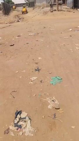 The video shows a dirt road in a village or town. Litter is scattered on the ground. A person is pushing a yellow cart toward the camera. Another person walks in the distance on the same road. Buildings are visible on either side of the road. Wires are suspended above the road.