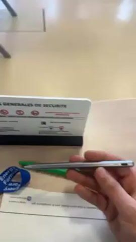 A hand spins a gray stylus between the fingers. It is then held in a vertical position and dropped onto a countertop. A sign describing safety regulations and an additional green pen are visible.