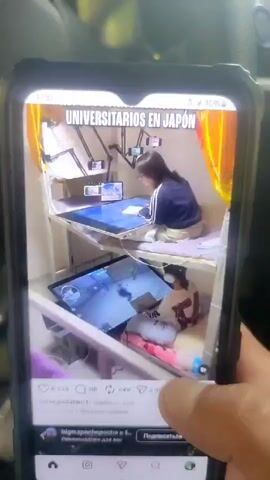 The video, presented on a smartphone, shows two different social media posts. The first post, in Spanish, displays a cramped living space, likely a dormitory room in Japan, with two individuals engaged in activities at desks. The second post, in English, features a woman driving a vehicle, with text overlay indicating "wait for it..." The video appears to be filmed by someone holding the phone, capturing the screen content.