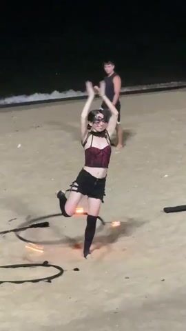 A person is doing a fire dancing performance on a beach at night. The person is wearing a red and black corset, black shorts, and black thigh-high socks. They have a black mask on their face and their arms are raised in the air. There is a hoop with fire on it around their waist, and two burning torches are on the sand to the left and right of them. The ocean is visible in the background. Another person is standing in the background watching the performance.
