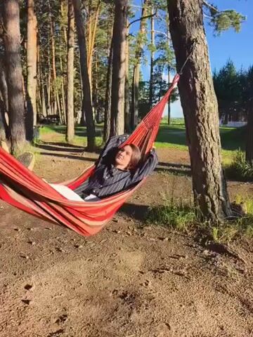 A woman lies in a striped hammock strung between two trees in a wooded area. She is wearing a dark shirt and white pants. Sunlight filters through the trees, casting shadows on the ground. The woman looks to her right at the camera then closes her eyes. The hammock sways gently.