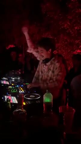 The video shows a DJ performing at an outdoor party at night, which has a red light illuminating everything. The DJ is male, with dark hair and a mustache, wearing a patterned shirt and headphones. He is operating a complex DJ setup with multiple turntables and control panels, manipulating the music with his hands and gesturing to the crowd with his fingers. Behind him, a group of people are gathered, some obscured by darkness but silhouettes of their faces are visible. The foreground features a table cluttered with bottles of liquor, drinks in plastic cups, and other miscellaneous items. The overall atmosphere is that of an active, vibrant outdoor party with music and socializing, where drinks are available.