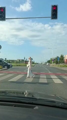 A video captures a woman in a white polka dot dress crossing a pedestrian crosswalk. Traffic lights are red, indicating that vehicles must stop. A black SUV is visible behind the woman, stopped at the red light. The scene takes place on a sunny day with a clear blue sky and some clouds. The road appears to be a multi-lane street in a suburban or urban area, with buildings and trees visible in the background. The perspective of the video is from inside a vehicle, looking forward through the windshield.