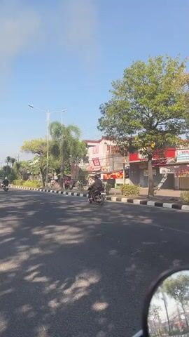 A vertical, medium-range shot captures traffic moving along a paved road on a bright, sunny day. The road is lined with trees and buildings. In the foreground, the side-view mirror of a vehicle is visible at the bottom of the frame. Several motorcycles are present in the video. One red and white motorcycle with a rider wearing a helmet and backpack is in the right lane. Further down the road, a person on a black motorcycle with a pink helmet is visible. In the left lane, a person on a white scooter with a blue shirt and helmet is seen, followed by another motorcycle. The sky is clear and blue with some wispy clouds. The scene appears to be in a town or city with businesses visible along the roadside.