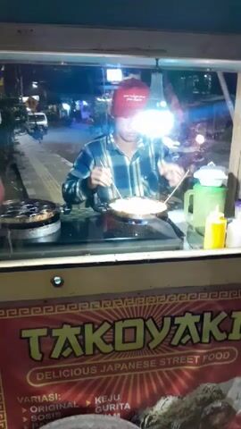 A man wearing a plaid shirt and a red baseball cap is cooking takoyaki at a street food stall at night. He is using chopsticks to turn the takoyaki balls in a specialized pan. A bright light bulb hangs above him, illuminating the scene. The stall has a sign that reads "TAKOYAKI DELICIOUS JAPANESE STREET FOOD". There are bottles of condiments and other cooking equipment visible on the counter. The background shows a dimly lit street with vehicles and other stalls. Another person is partially visible in the background, possibly a customer.