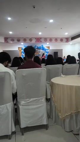 The video shows a scene indoors, likely a gathering or event. In the foreground, several people are seated facing away from the camera, their backs and the tops of their heads visible. White chair covers are visible. In the mid-ground, a stage area is decorated with blue and white balloons and silver tinsel curtains. Two women are on the stage. One, wearing a pink top, is dancing or performing. The other, partially obscured, appears to be a woman with long dark hair. A large speaker is visible on the right side of the stage. The environment appears to be a hall or banquet room with a tiled ceiling and lighting fixtures. A round table covered with a beige tablecloth is in the right foreground.