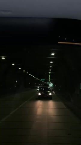 The video shows the view through a car's rear window as it travels through a dark tunnel. The tunnel is illuminated by rows of lights along the ceiling. Another car is visible ahead, its headlights illuminating the road. The car in the foreground appears to be moving at a moderate speed. The tunnel walls are visible on either side, and the road surface is marked with white lines. There are some green signs visible on the tunnel walls in the distance.