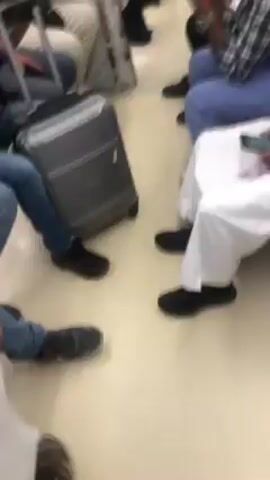 The video shows the interior of what appears to be a train car. Several people are sitting, some looking at their phones. A gray suitcase sits on the floor. Some of the people are wearing casual clothes like jeans and t-shirts, while one man is wearing a traditional white thobe. The lighting is bright. The camera moves around slightly, capturing different seated passengers.