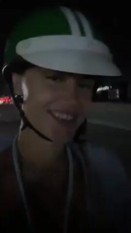 A woman is on a moped at night. She is wearing a green and white helmet and a light-colored tank top. She is smiling and then winks at the camera. She is wearing a necklace and small hoop earrings. You can see the traffic lights in the background.