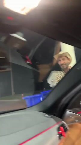 A man wearing a cowboy hat and a leopard-print shirt is standing next to a car, giving a thumbs up. He appears to be at a gas station, with a gas pump visible in the background. The video is shot from inside another car, looking out through the windshield. The man smiles and gestures with his hands as he speaks. There is a blue bucket on the ground near him.