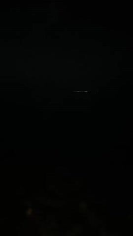 The video begins with a dark, seemingly water-filled environment. A faint horizontal line of light, possibly from distant structures or a boat, is visible in the upper portion of the frame. As the video progresses, the camera angle shifts downwards, revealing a dark, rocky shoreline or seabed. The water gently laps against the stones. The overall scene suggests a dark, possibly nighttime, outdoor setting with a body of water.