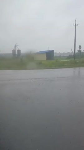 The video shows a road with a blurry foreground, indicating it was likely filmed through a wet window or lens. The background features a rural landscape under a heavy, overcast sky, suggesting rain or fog. There are several structures visible, including silos, smaller buildings, and utility poles. The ground appears wet, and the overall atmosphere is dim and hazy.