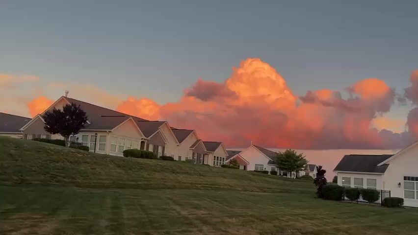The video shows a row of identical ranch-style houses with manicured lawns and vibrant pink and orange clouds in the sky. The houses are white with brick accents and have attached garages. A black asphalt driveway leads to the garages, and a small American flag is visible in the foreground. The sky is clear except for the large, dramatic clouds that are illuminated by the setting sun. The overall scene is serene and picturesque, conveying a sense of suburban tranquility.