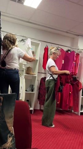 The video shows two women browsing in a clothing store with a red carpet. The woman in the foreground wears a white and gray striped t-shirt and green pants, and examines a red top. The woman in the background wears a white t-shirt and jeans and looks at items on a shelf. Clothing racks with pink, red, and other colored clothes, as well as a white shelf displaying shoes, are also visible.