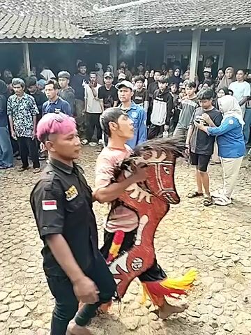 The video shows an outdoor event in front of a building. In the foreground, two individuals are interacting with a large, colorful prop that resembles a horse. One person, wearing a black shirt with an Indonesian flag patch and pink hair, appears to be guiding or holding the prop. The other person, in a light-colored, patterned shirt, is actively manipulating the prop. Both are dressed casually. A crowd of spectators, including men, women, and children, are gathered behind them, watching the activity. The environment appears to be a village or rural setting, with a dirt or gravel ground and traditional-looking architecture. The overall atmosphere seems to be one of cultural performance or celebration.