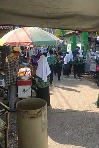 A vertical video shows an outdoor scene, likely a street or walkway, with a group of people, mostly young students, wearing green uniforms and headscarves. They appear to be in a procession or lining up. One person in the foreground, a man wearing a plaid shirt and a hat, is facing away from the camera, near a food cart. The environment includes pavement, shadows from sunlight, and some greenery and buildings in the background. The camera seems to be moving slightly, creating a subtle blur.