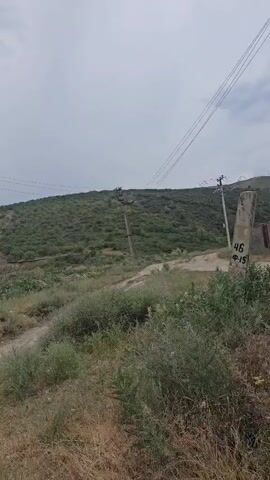 The video shows a person walking on a dirt path towards a road. The person is wearing a white harness and dark pants. There are several cars parked along the side of the road, and a few people are standing near the cars. The road appears to be in a rural or semi-rural area, with hills covered in vegetation in the background. The sky is overcast.