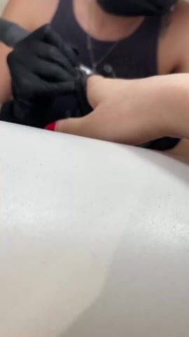 Here's a detailed description of the video:

A manicurist, wearing black gloves and a black tank top, is seen using an electric file on a person's fingernail. The manicurist also has a black face mask on. The person whose nail is being filed is resting their hand on a white surface. The fingernail has red nail polish on it. The manicurist's hands are moving deliberately, and the electric file is making a buzzing sound. The setting appears to be a nail salon.
