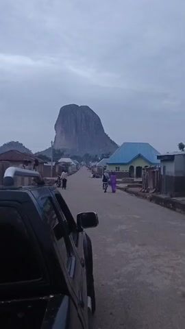 The scene shows a straight paved road through a rural village, with a large, prominent rock formation in the distance. The rock has a distinctive rounded shape with visible vertical striations, appearing monolithic and dominating the skyline. On either side of the road are simple, low-lying buildings, some with corrugated metal roofs and others with painted surfaces. People are walking along the road, some in traditional attire. A black pickup truck is parked on the left side of the road, partially obstructing the view. The sky is overcast, suggesting an early morning or late afternoon setting. The lighting is muted and even, emphasizing the gray tones of the landscape and the rock formation.