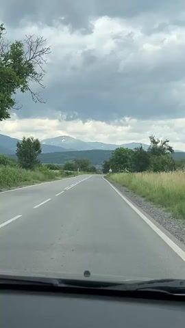 The video shows a car driving down a country road on a cloudy day. The road has two lanes and white lines, with grass and trees on either side. In the distance, there are mountains, some of which have snow on top. There is a road sign indicating a curve ahead. The footage is taken from the dashboard of the car.