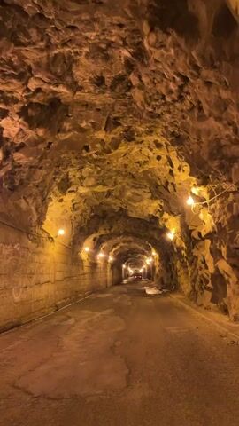 The video shows the inside of a long tunnel. The walls and ceiling are rough and rocky, lit by evenly spaced lights. The road is cracked and uneven. There is a car visible in the distance at the end of the tunnel.
