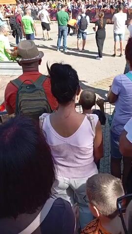 The video shows a crowded outdoor event with people watching something happening in the distance. In the foreground, several people are viewed from behind, suggesting they are spectators. One person in the middle ground wears a hat and a backpack. The background is filled with a larger crowd and individuals walking around. The setting appears to be a public space, possibly a street or square, with some temporary structures or barriers visible. The overall atmosphere seems lively, with an announcer possibly speaking, indicated by the partial speech heard.