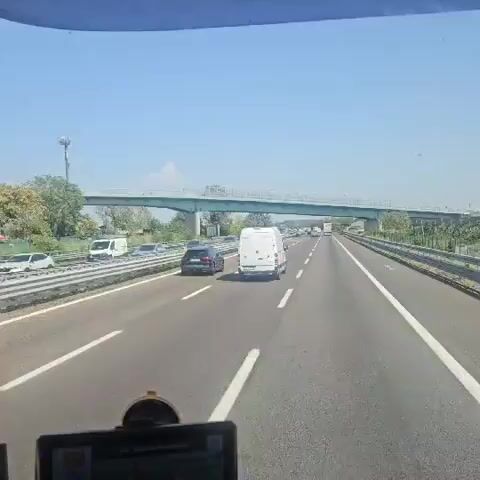 The video shows a view from inside a vehicle traveling on a highway. A green overpass is visible in the distance, with cars and trucks driving under it. The highway is multi-lane, with white lane markings. The sky is clear and blue. The video appears to be taken during the daytime.