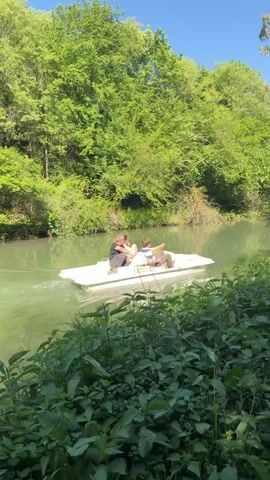 A pedal boat carrying four people moves slowly down a narrow, green river. A woman with blonde hair, sunglasses, and a pink shirt walks on the riverbank. The river is surrounded by dense green trees and foliage. The sky is bright blue.