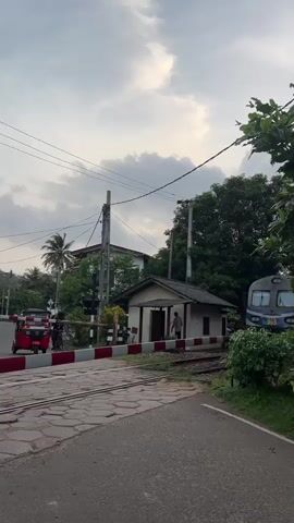 A blue and white train, numbered 995, passes through a railroad crossing. The crossing gate is down. A red tuk-tuk is stopped at the crossing. There are power lines and trees in the background. The sky is cloudy.