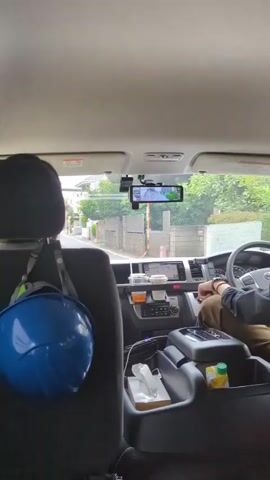 This is a point-of-view video from inside a vehicle, likely a van, looking forward. The camera is positioned behind the front passenger seat, with a blue helmet hanging from the headrest in the foreground. The driver is visible in the driver's seat, operating the vehicle. The dashboard displays a navigation screen and other vehicle controls. The road ahead is visible through the windshield, with houses and trees lining the street. The video captures the movement of the vehicle as it drives down the road.