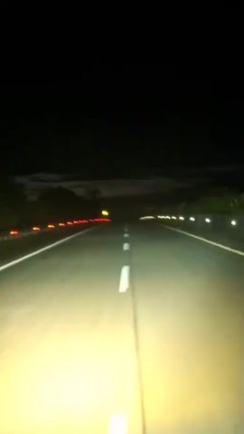 The video shows a low-angle perspective from the front of a vehicle driving on a dark road at night. The headlights illuminate the asphalt, revealing a white solid line in the center and a white dashed line on the right side. Red reflective markers line the left shoulder of the road, while white reflective markers appear on the right shoulder. A yellow road sign is visible in the distance. The sky is dark with some clouds. An unknown melodic music plays in the background.