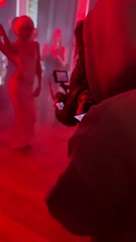 The video shows a group of people on a wooden floor, illuminated by red lighting and fog. The focus is on the backs of several people, including one wearing a black jacket with curly hair and another in a light brown hoodie. In the background, there are more individuals, some in casual clothing, and a person holding a professional-looking video camera. The atmosphere suggests a filming event or party, with the camera capturing the scene while people mill around.