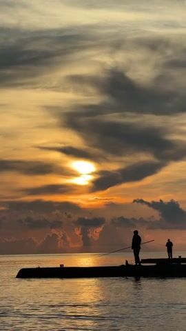 The video shows a sunset over the ocean. Two people are fishing from a pier. The sun is low in the sky, casting a warm orange glow. Clouds are visible in the sky, and the water is calm. The silhouettes of the two fishermen are clearly visible against the bright sky.