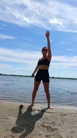 A woman in a black sports bra and black shorts stands on a sandy beach with her arms extended. She raises her right arm straight up, then lowers it and raises her left arm. She then brings both arms up in front of her and moves them out to her sides. The ocean is behind her, and the sky is blue with scattered clouds.