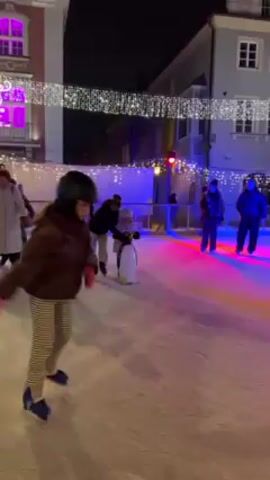 The video shows a winter ice rink at night. Several people are ice skating, including adults and children, under strings of lights strung between buildings. Some children are using penguin-shaped skate aids to help them balance. The ice is illuminated with colored lights, creating a festive atmosphere. The buildings surrounding the ice rink are visible in the background. The camera appears to be handheld, and the perspective shifts as the camera operator moves around the rink.