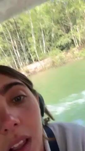 The video shows a woman taking a selfie on a moving boat. She is looking into the camera while the boat moves on the water. Behind her is the green shore with trees. The boat has a roof. The water is green and churning from the boat's movement. Another person's shoulder and head are briefly visible beside the woman.